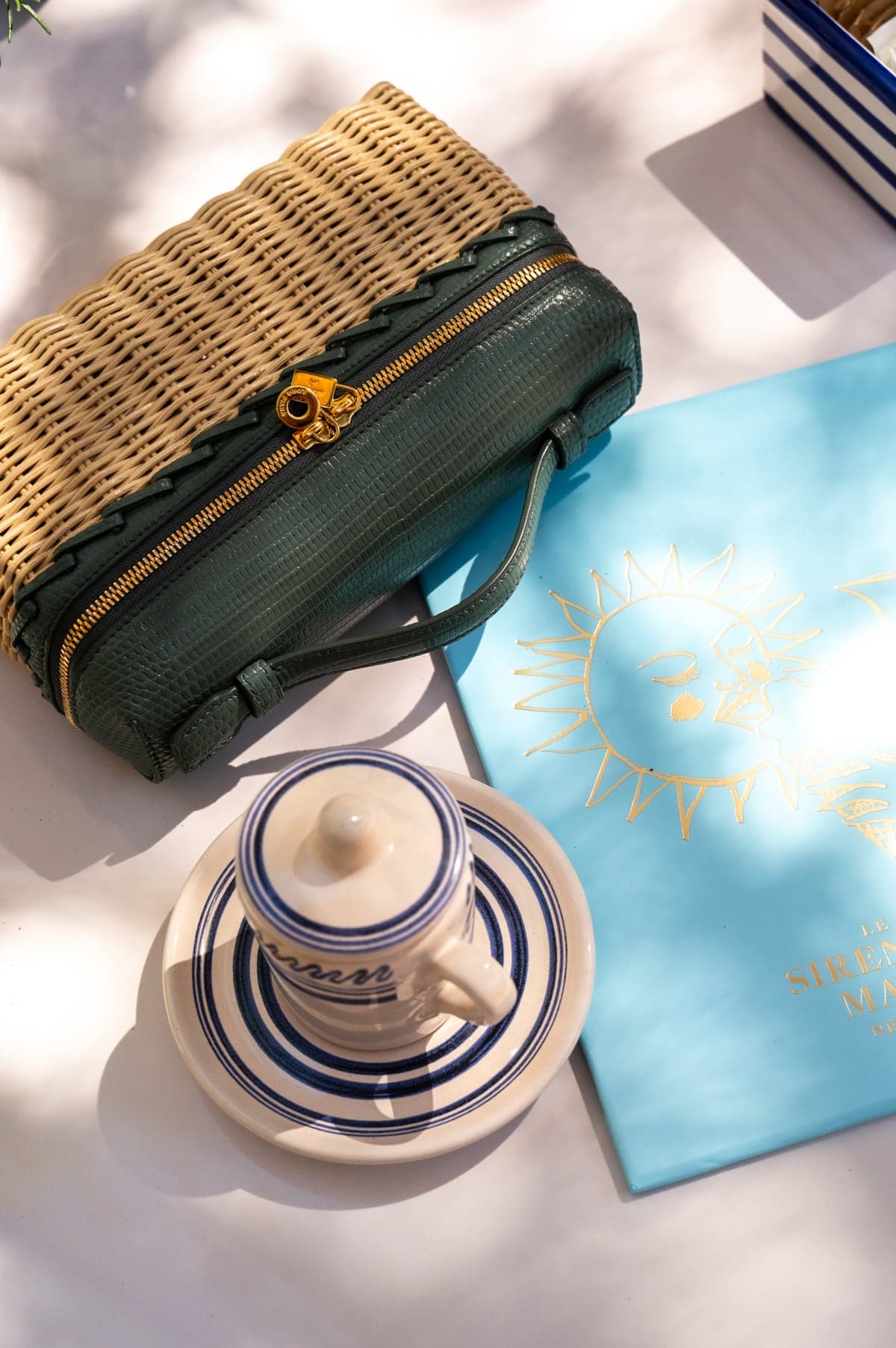 A wicker-and-green-leather clutch beside an espresso cup and a blue Sirenuse menu in the sun